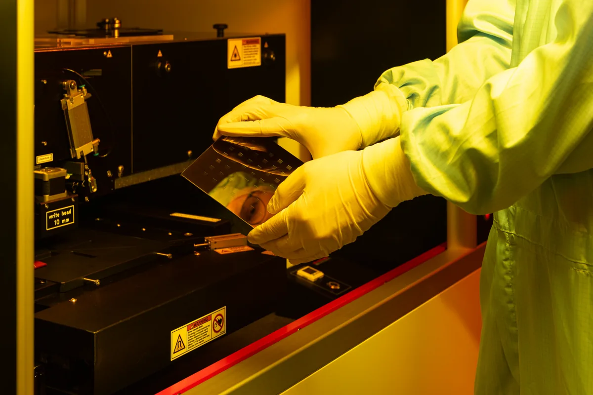A researcher in cleanroom clothing holds a patterned wafer beside laser lithography equipment.